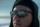 A close-up of a man wearing sunglasses and a hat, looking at something off camera. A road is seen reflected in his sunglasses.